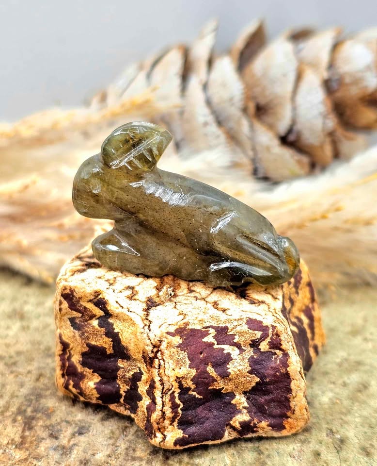 One hand carved rabbit figurines displayed on a natural wood slice, with a pinecone and dried grass in the background.