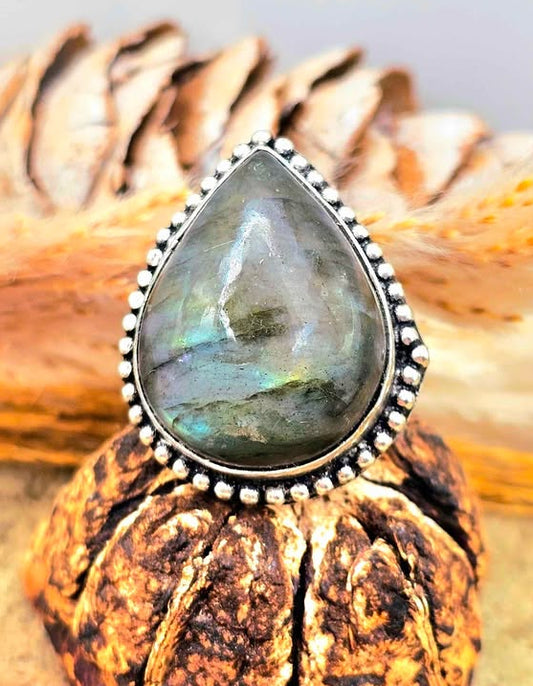 A teardrop-shaped labradorite ring with a silver beaded border, displayed on a textured brown seed pod with dried grass and pine cones in the background.