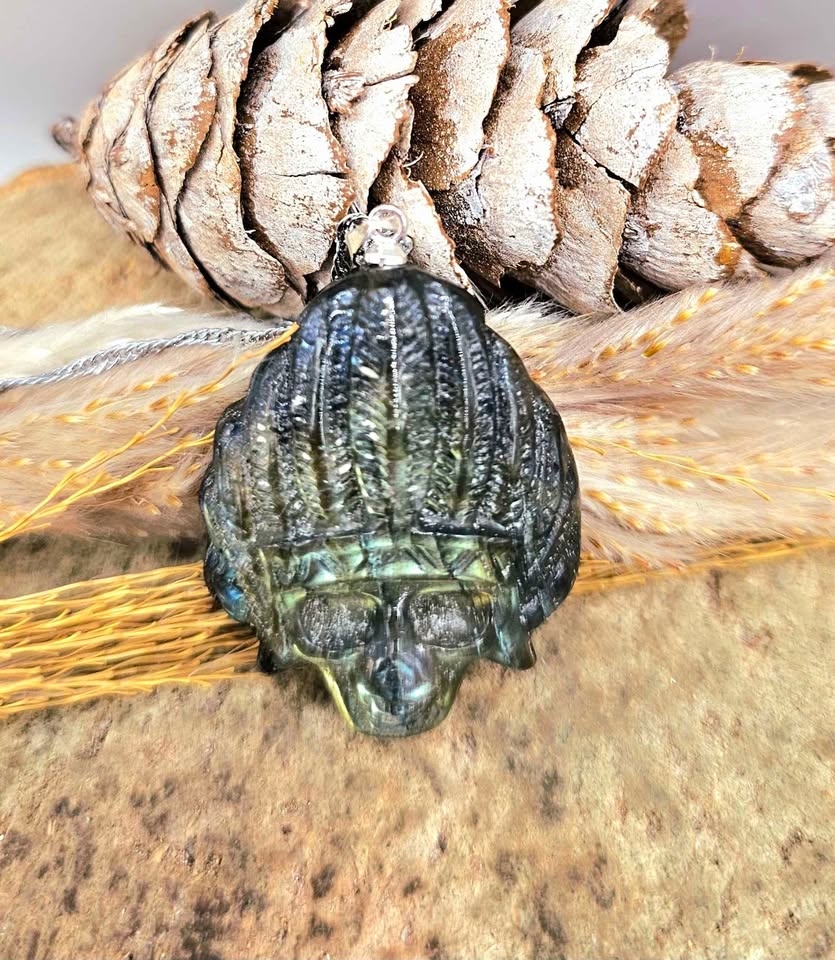 A dark metallic skull-shaped pendant with a textured, ribbed surface, resting on a rustic background of a pinecone, dried wheat, and wood.