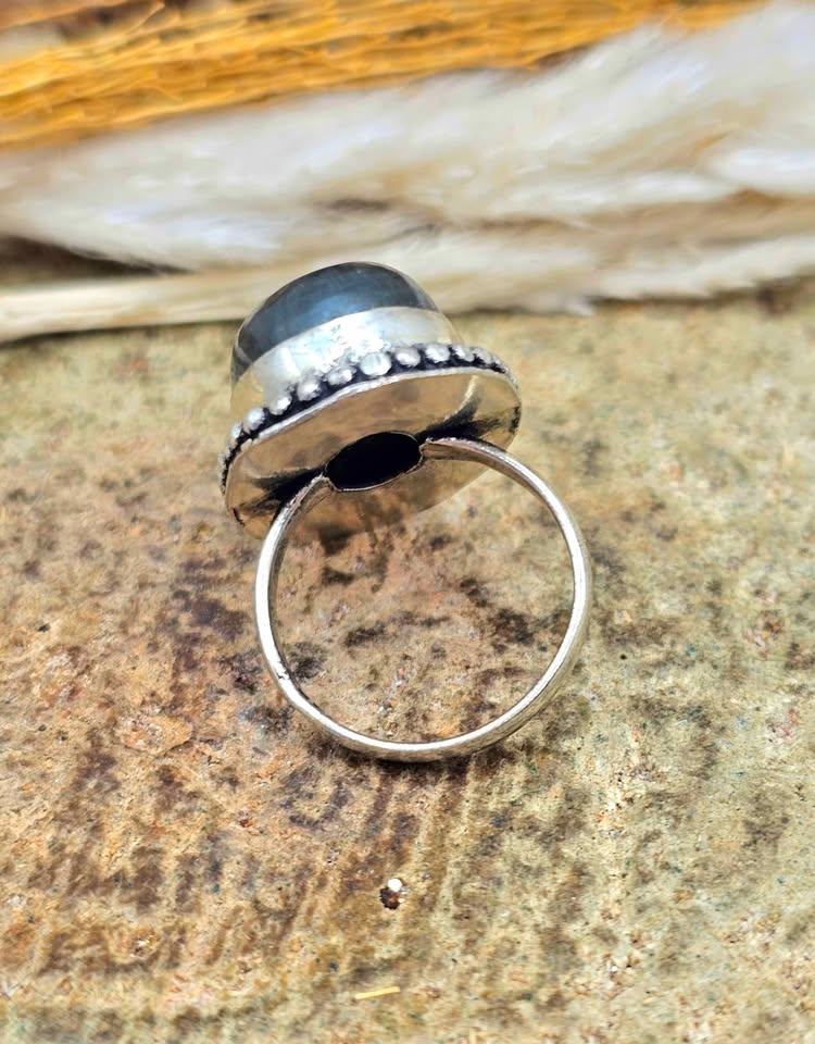 A sterling silver ring with a large round black stone, photographed close-up on a textured stone surface with dried grass in the background.