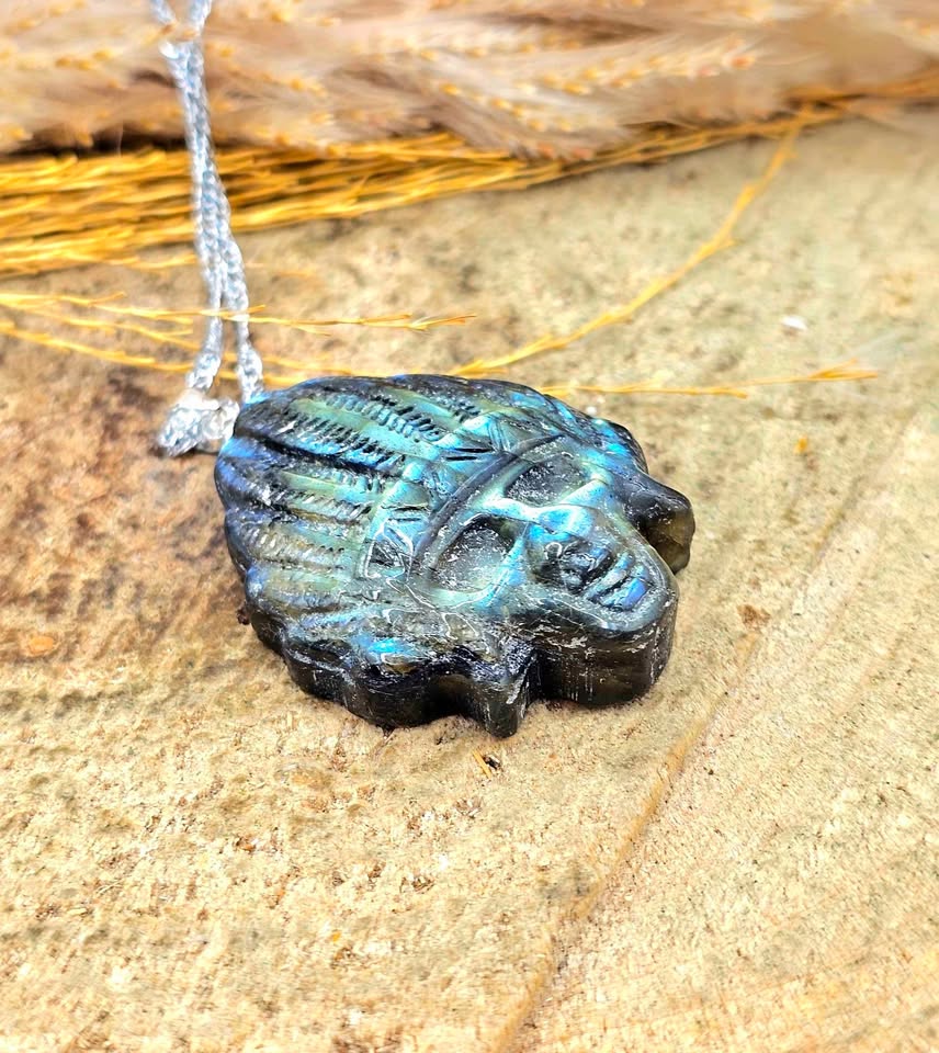 A carved black stone pendant with a blue-green iridescent patina, shaped like a stylized face or mask, resting on a textured tan stone surface with a silver cord visible at the top.