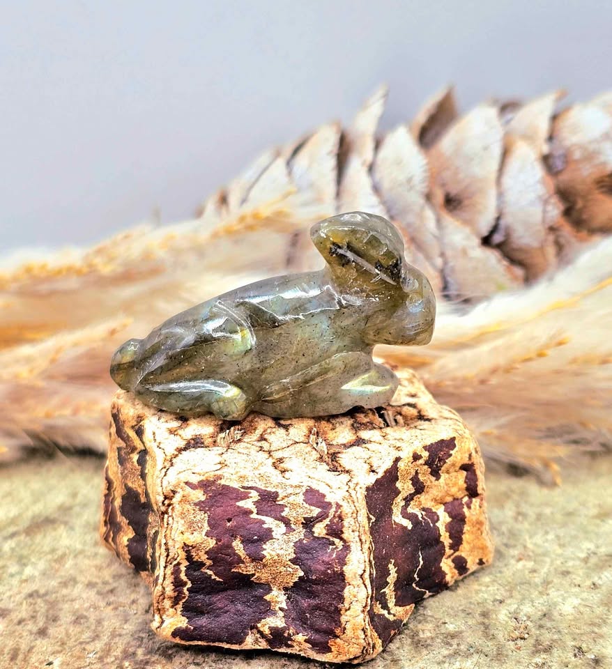 One  hand carved rabbit figurines displayed on a natural wood slice, with a pinecone and dried grass in the background.