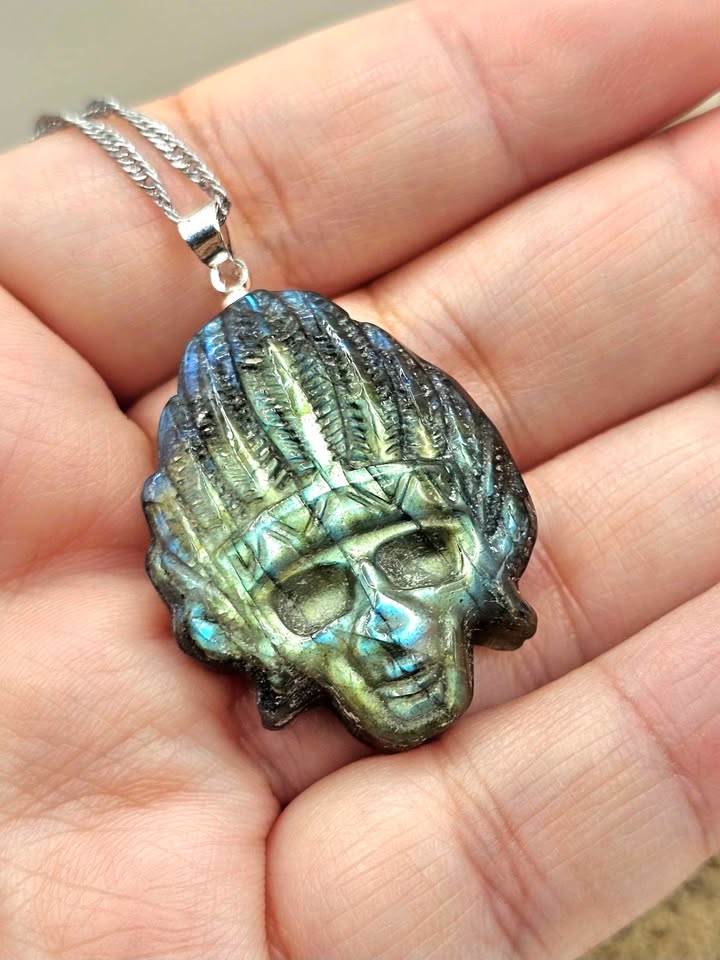 A close-up of a hand holding a silver pendant necklace featuring a detailed skull with textured hair and an iridescent blue-green labradorite inlay, attached to a silver chain.