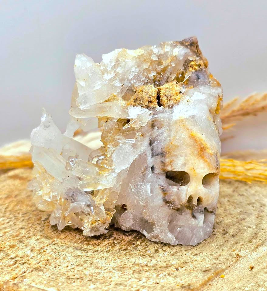 A natural crystal cluster featuring a prominent skull formation with clear quartz and beige mineral inclusions, displayed on a textured burlap surface-in closed up front view- Aura Gemstones. 
