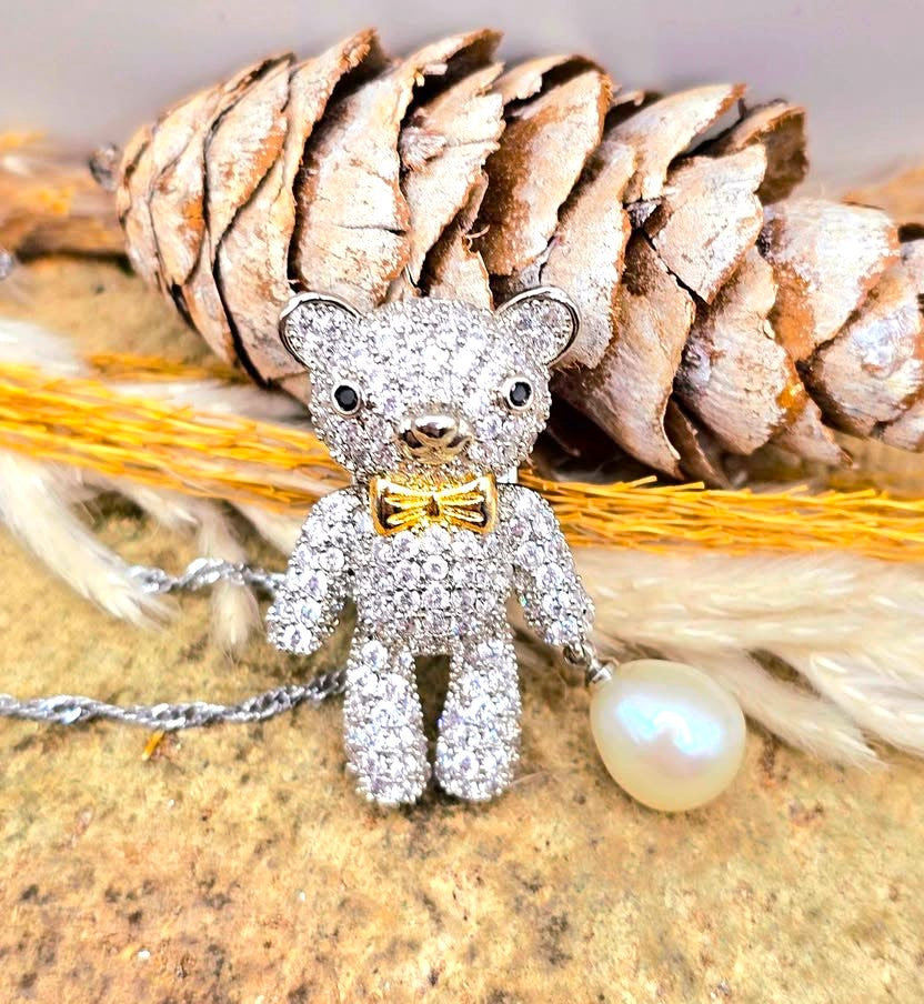 A teddy bear pendant covered in sparkling clear stones, wearing a gold bow tie, with a silver chain and a white pearl hanging from one hand in top view-My Aura Gemstones