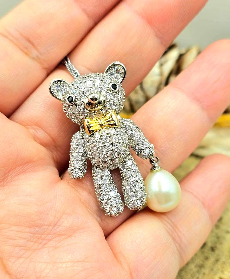 A teddy bear pendant covered in sparkling clear stones, wearing a gold bow tie, with a silver chain and a white pearl hanging from one hand and display on a hand-My Aura Gemstones