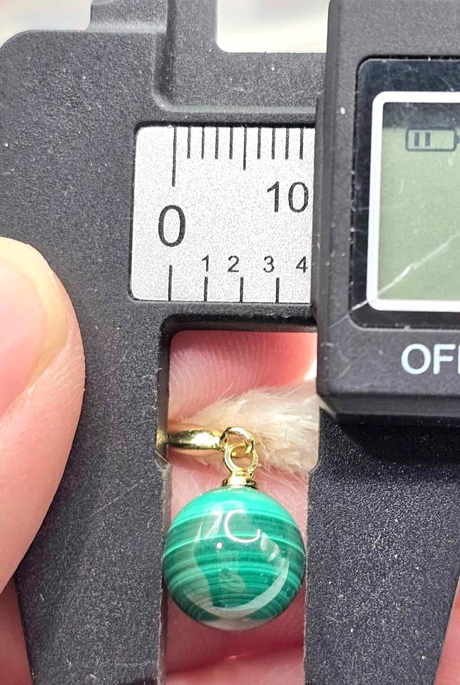 A close-up of a green and white banded malachite bead charm with a gold bail, shown being measured by a digital caliper held in a hand.