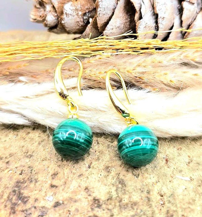A pair of gold-tone drop earrings with round green malachite beads, displayed on a textured surface with natural props like a pinecone and dried grass-My Aura Gemstones