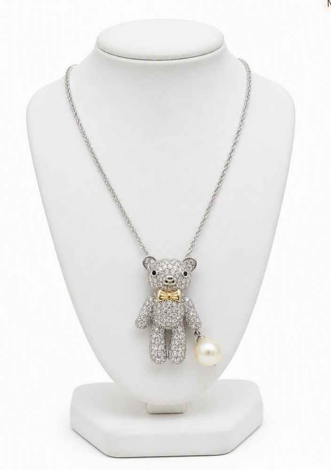 A teddy bear pendant covered in sparkling clear stones, wearing a gold bow tie, with a silver chain and a white pearl hanging from one hand and display in jewelry stand My Aura Gemstones