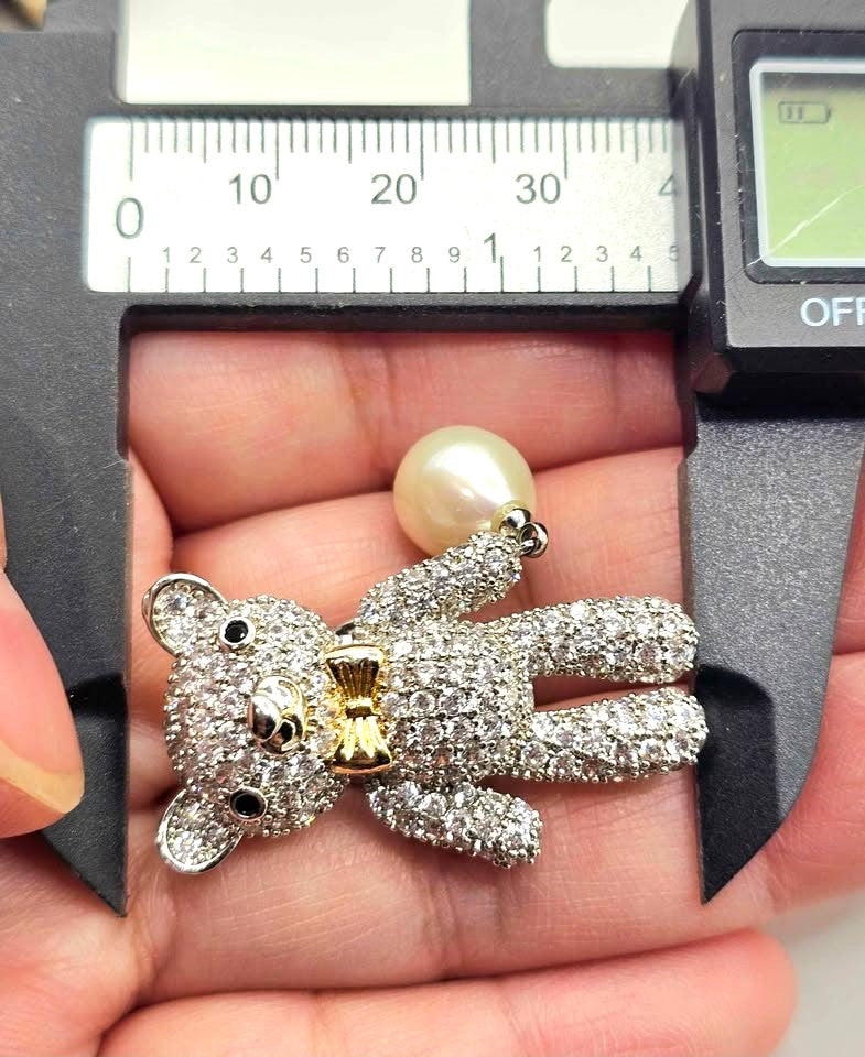 A teddy bear pendant covered in sparkling clear stones, wearing a gold bow tie, with a silver chain and a white pearl hanging from one hand with measurement -My Aura Gemstones