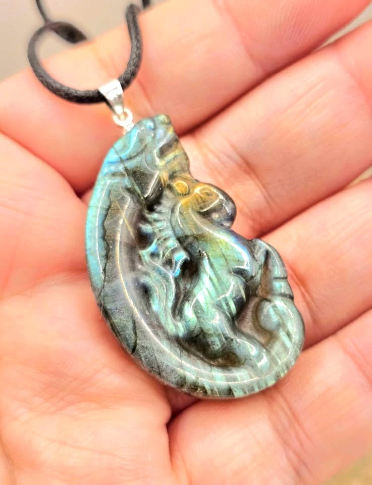 A carved labradorite  horse with a dragon pendant necklace featuring a detailed figure design with a shimmering blue-green iridescent on a hand- My Aura Gemstones. 