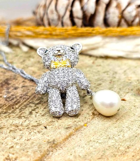 A teddy bear pendant covered in sparkling clear stones, wearing a gold bow tie, with a silver chain and a white pearl hanging from one hand in flat view-My Aura Gemstones