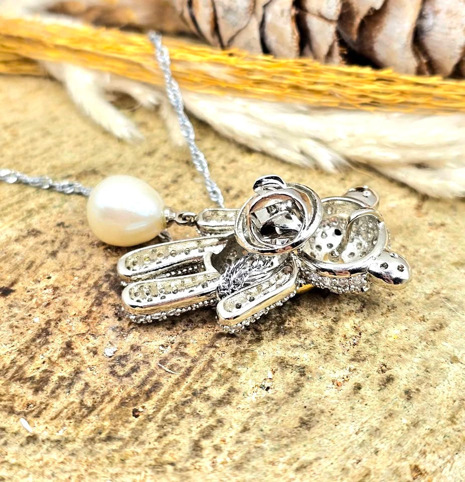 A teddy bear pendant covered in sparkling clear stones, wearing a gold bow tie, with a silver chain and a white pearl hanging from one hand in back view-My Aura Gemstones