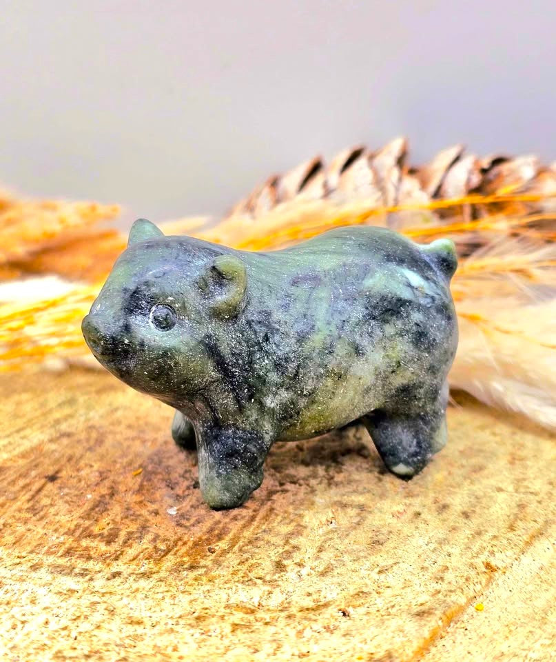 A small Serpentine carved animal figurine, shaped like a pig or boar, made from dark green stone with natural speckling, standing on a textured wooden surface with dried grass and a pinecone in the background - My Aura Gemstones