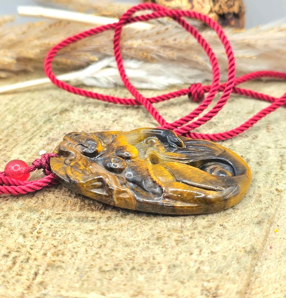 A carved oval pendant made of golden-brown tiger's eye stone carved mermaid,  pendant threaded on a red cord with a red bead in another side view-My Aura Gemstones