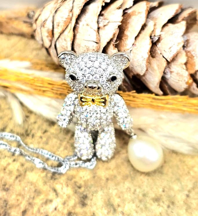 A teddy bear pendant covered in sparkling clear stones, wearing a gold bow tie, with a silver chain and a white pearl hanging from one hand in top view-My Aura Gemstones