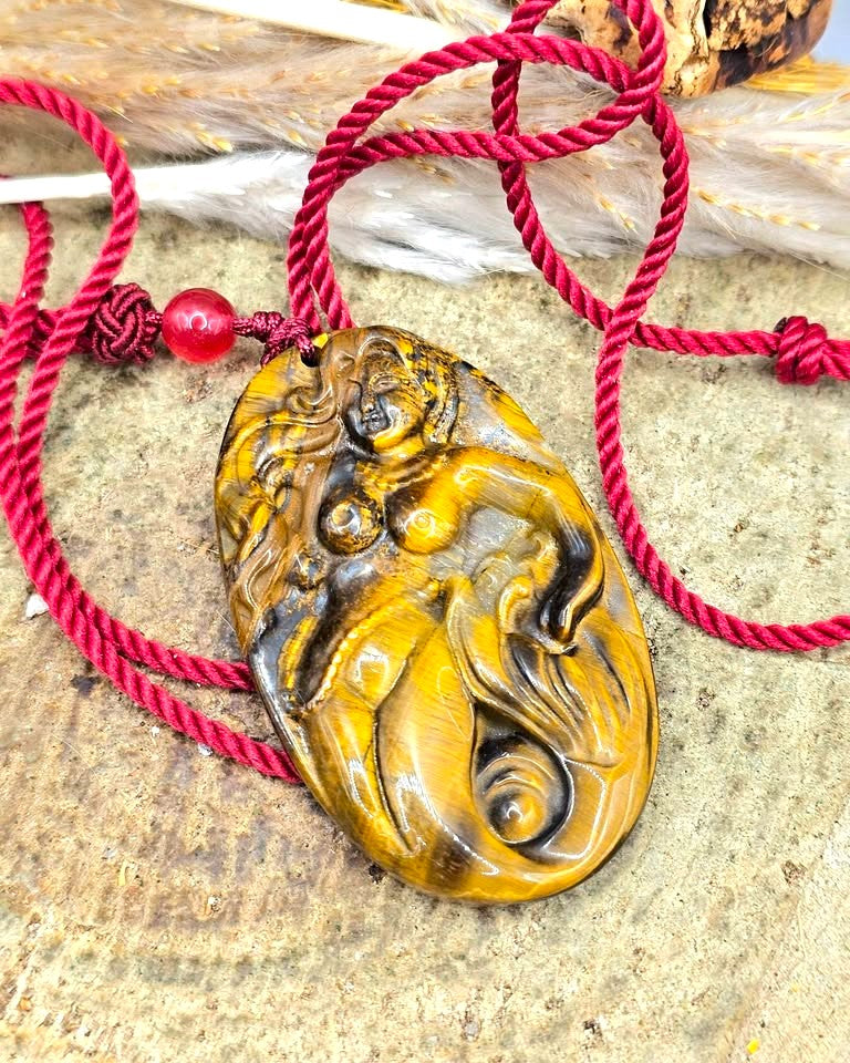 A carved oval pendant made of golden-brown tiger's eye stone carved mermaid,  pendant threaded on a red cord with a red bead in top view-My Aura Gemstones