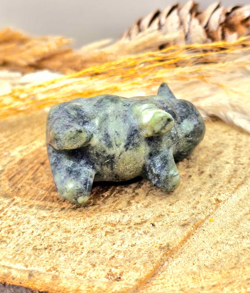 A small Serpentine  stone figurine of a sleeping animal, likely a rabbit or hare, in green and gray marbled stone, displayed on a textured tan surface with dried grass and a pinecone in the background.
