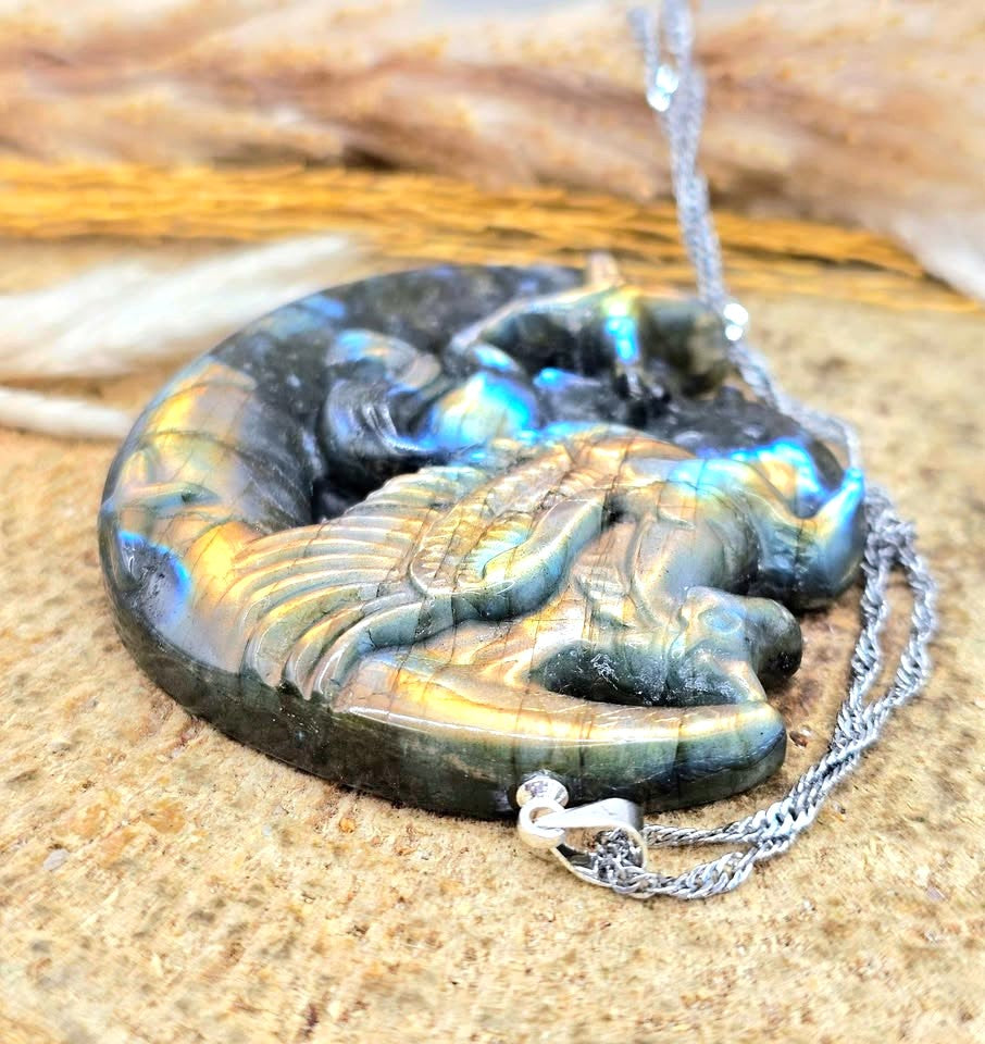 A carved labradorite pendant featuring a winged horse (Pegasus) resting on a crescent moon, displayed on a textured stone surface with dried grass in bottom view-My Aura Gemstones
