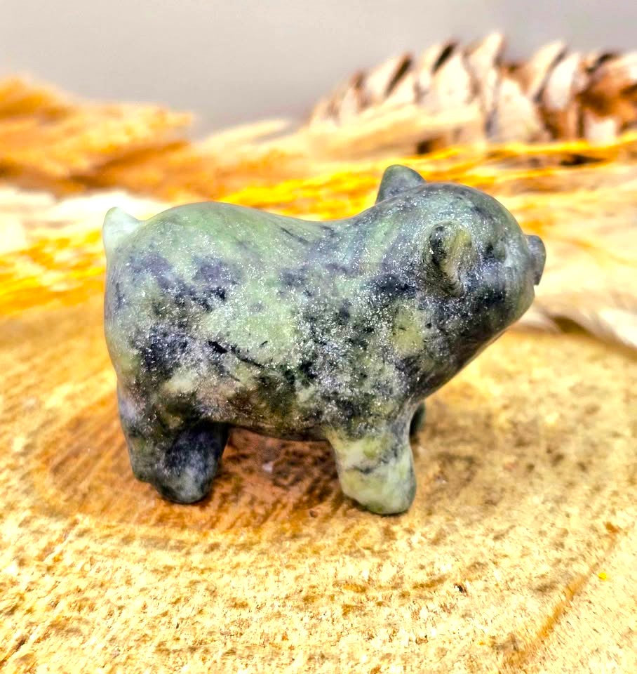 A small Serpentine carved animal figurine resembling a pig or boar, made from a speckled green and dark gray stone, standing on a textured wood slice with dried leaves in the background.