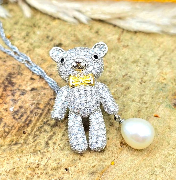 A teddy bear pendant covered in sparkling clear stones, wearing a gold bow tie, with a silver chain and a white pearl hanging from one hand at top view-My Aura Gemstones