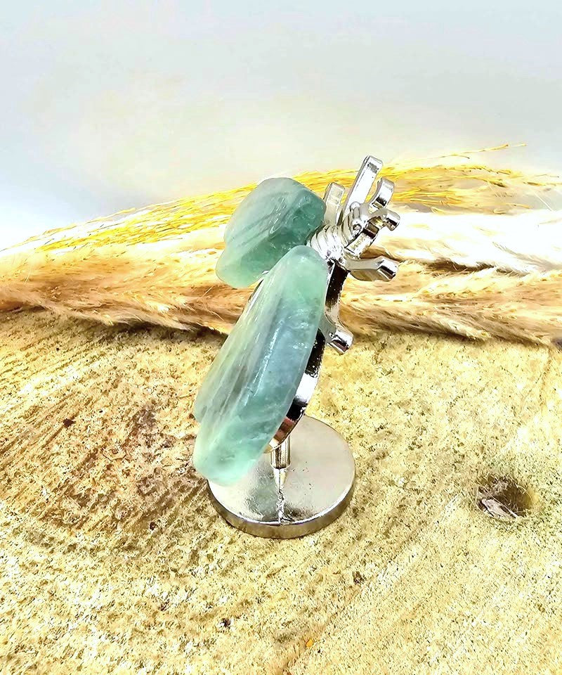 A silver metal bee figurine with natural fluorite wings, mounted on a round silver base and displayed on a textured sandy surface at another in another side view- My Aura Gemstones