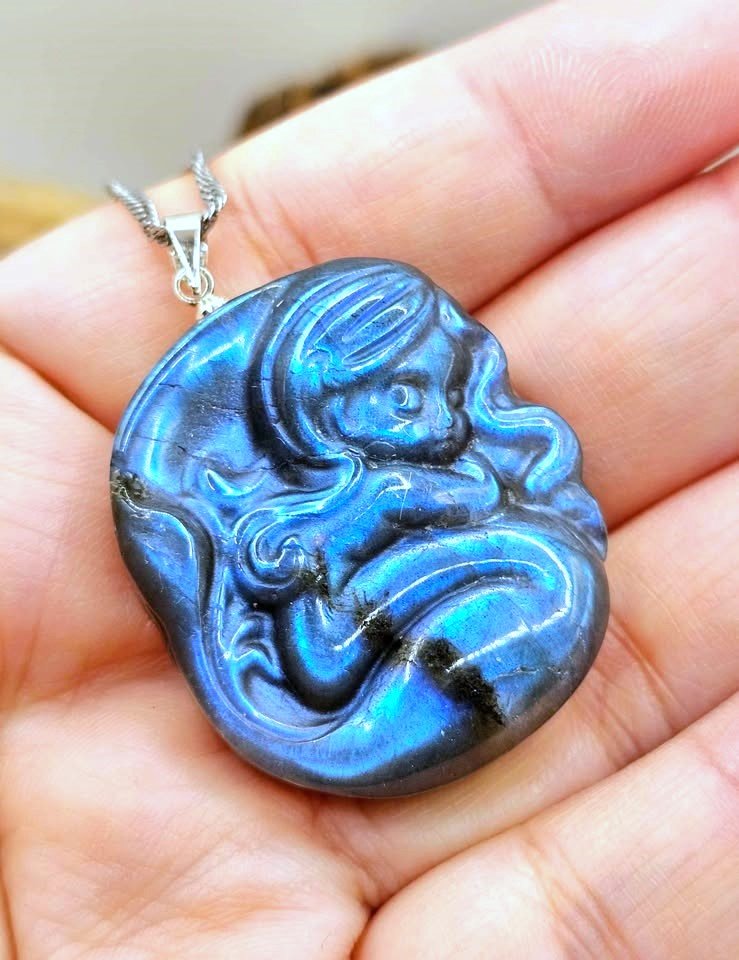 A round iridescent blue flash labradorite carved pendant with a carved fantasy sleeping mermaid design, resting on hand-My Aura Gemstones