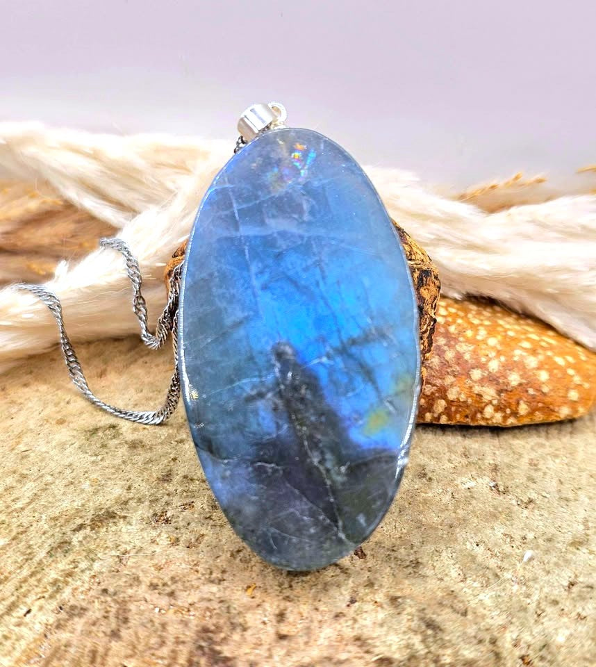 Labradorite carved pendant featuring a mermaid design, suspended from a silver chain, displayed on a sandy surface with dried grass and a small shell in the background in back view -My Aura Gemstones