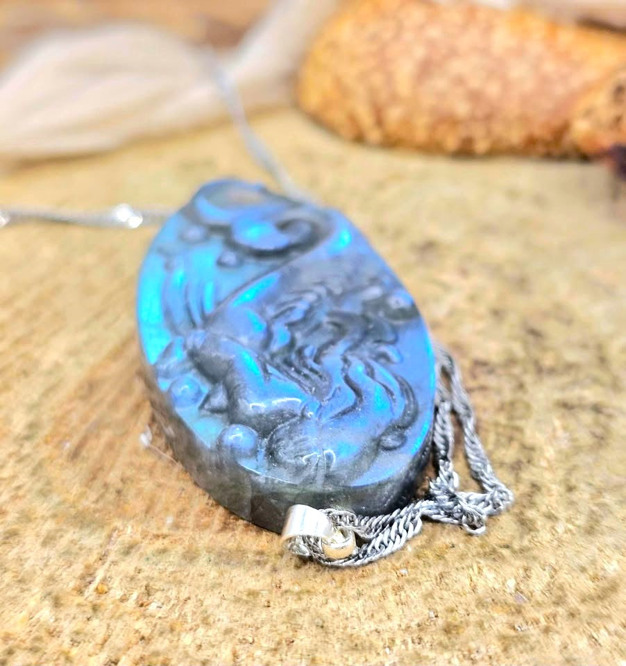 Labradorite carved pendant featuring a mermaid design, suspended from a silver chain, displayed on a sandy surface with dried grass and a small shell in the background in top view-My Aura Gemstones