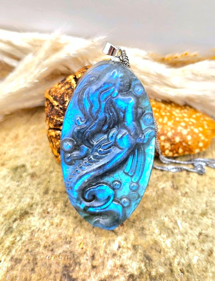Labradorite carved pendant featuring a mermaid design, suspended from a silver chain, displayed on a sandy surface with dried grass and a small shell in the background in front view-My Aura Gemstones
