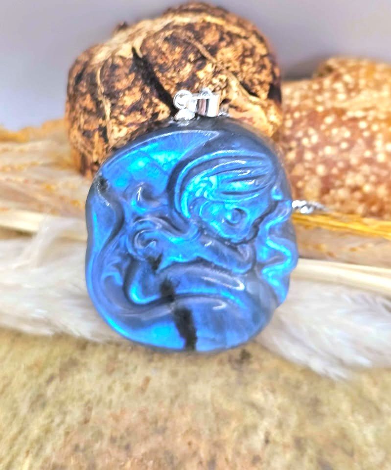 A round iridescent blue flash labradorite carved pendant with a carved fantasy sleeping mermaid design, resting on a textured stone surface with white feathers and dried grass in the background in another top view -My Aura Gemstones