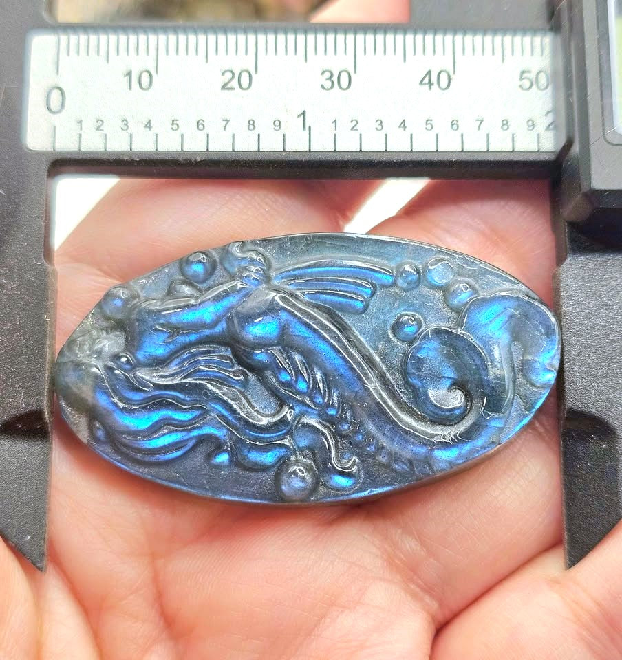 An oval carved stone pendant with a mermaid and wave design, held in a hand under a metal caliper with a ruler visible. The stone shows a strong blue iridescent labradorite effect.