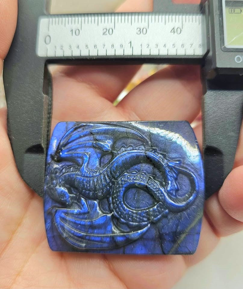 A hand holds a carved blue labradorite stone pendant featuring a dragon design, with a digital caliper measuring its width above it.