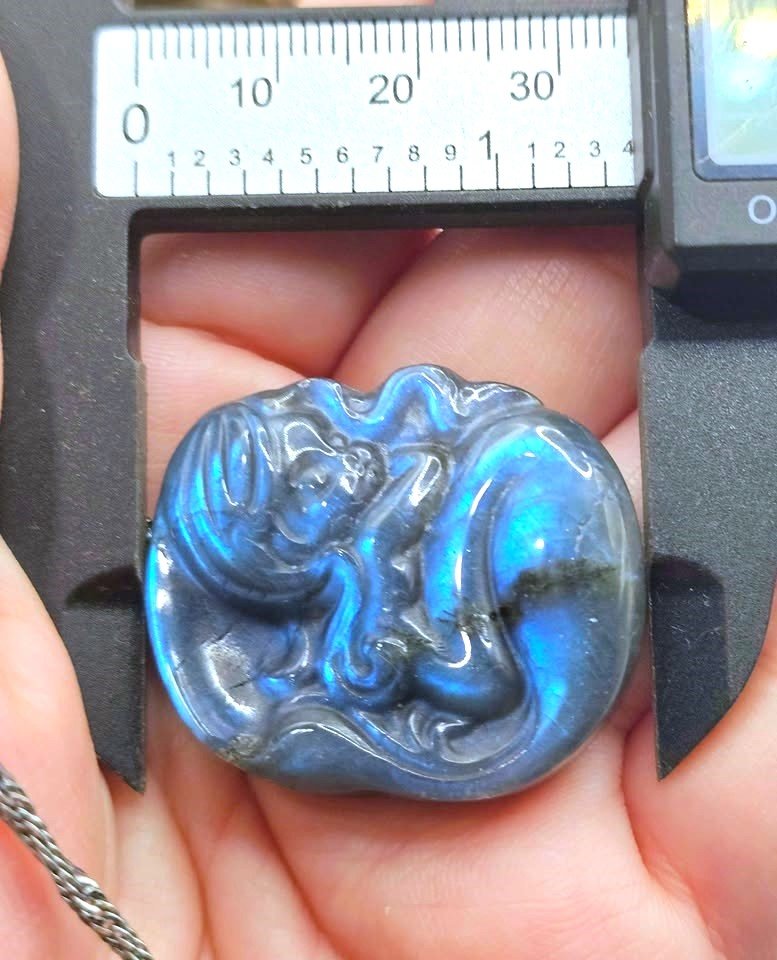A close-up of a carved blue iridescent stone pendant held in a hand under a caliper for scale. The pendant has a glossy, polished finish and features swirling, cloud-like patterns with strong blue and purple hues.