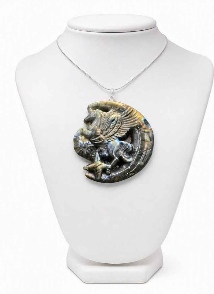 A carved labradorite pendant featuring a winged horse (Pegasus) resting on a crescent moon, displayed on white jewelry stand-My Aura Gemstones.

