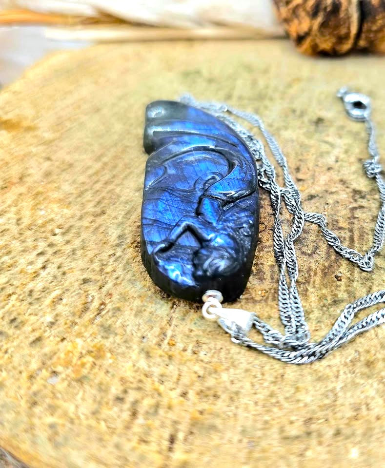 A carved labradorite mermaid pendant necklace featuring a stylized figure design, resting on a textured wooden surface. The pendant exhibits a vibrant blue and black iridescent labradorite finish, suspended from a silver-toned chain.