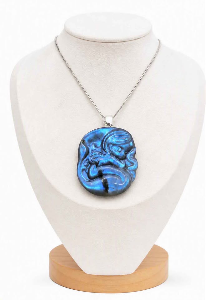 A round iridescent blue flash labradorite carved pendant with a carved fantasy sleeping mermaid design, resting on a textured stone surface with white feathers and dried grass in the background display in jewelry stand-My Aura Gemstones