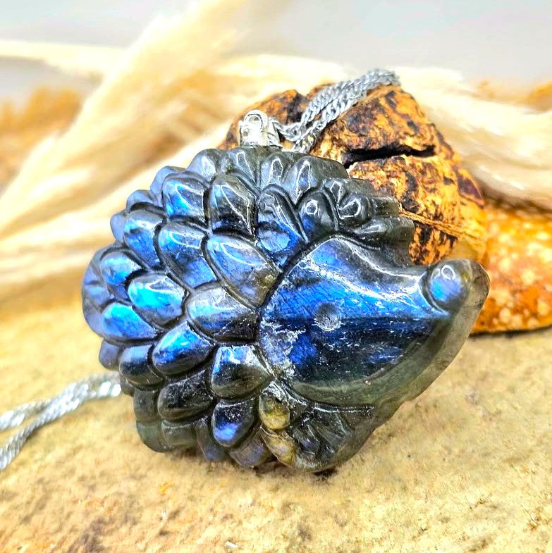 A carved labradorite hedgehog pendant with a silver bail, resting on a textured stone surface with dried grass in the background at another side view-My Aura Gemstones