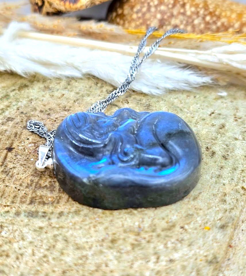 A round iridescent blue flash labradorite carved pendant with a carved fantasy sleeping mermaid design, resting on a textured stone surface with white feathers and dried grass in the background in side view-My Aura Gemstones