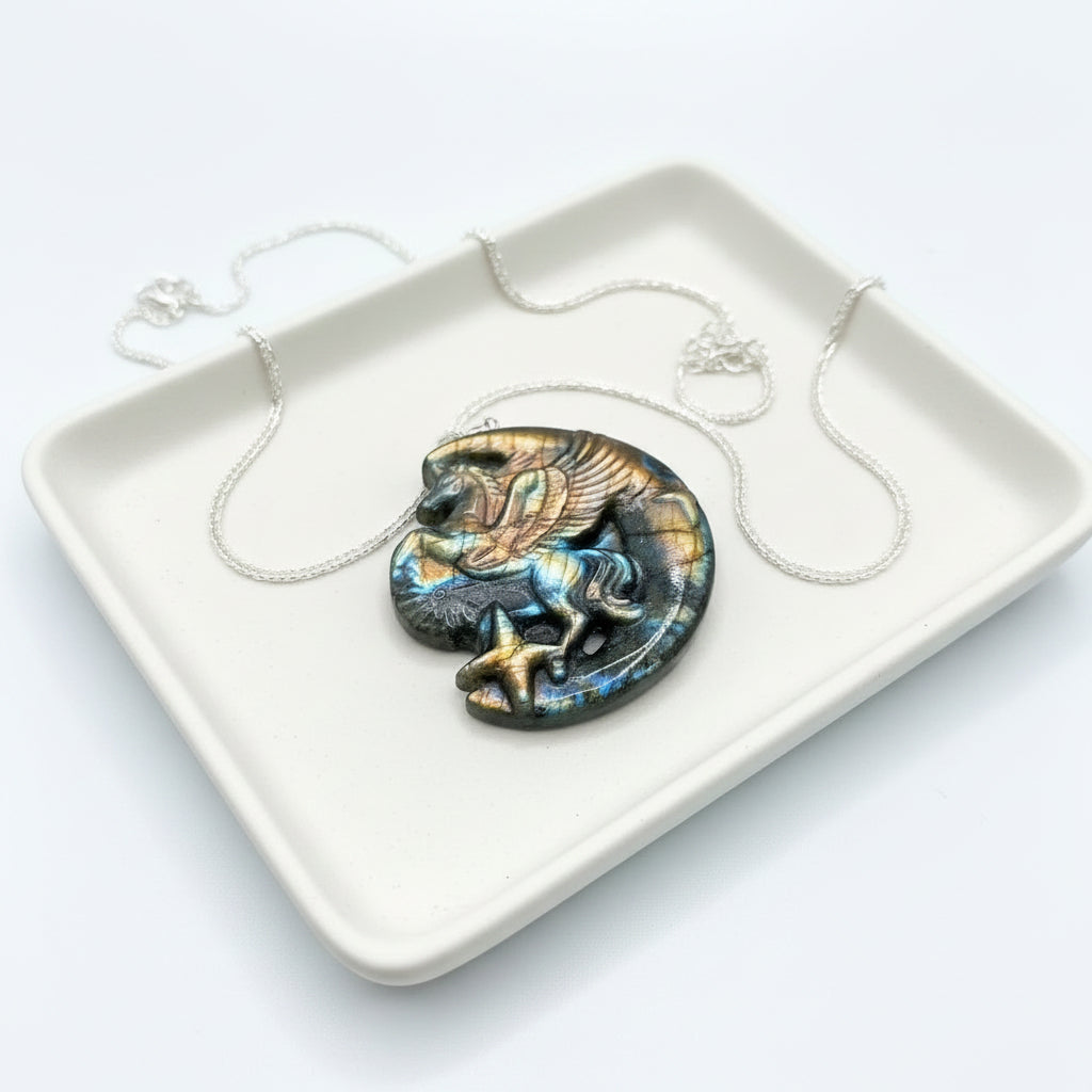 A carved labradorite pendant featuring a winged horse (Pegasus) resting on a crescent moon, displayed on a textured stone surface with dried grass-My Aura Gemstones
