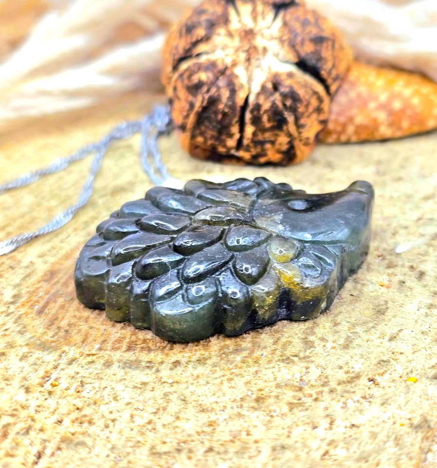 A carved labradorite hedgehog pendant with a silver bail, resting on a textured stone surface with dried grass in the background-My Aura Gemstones