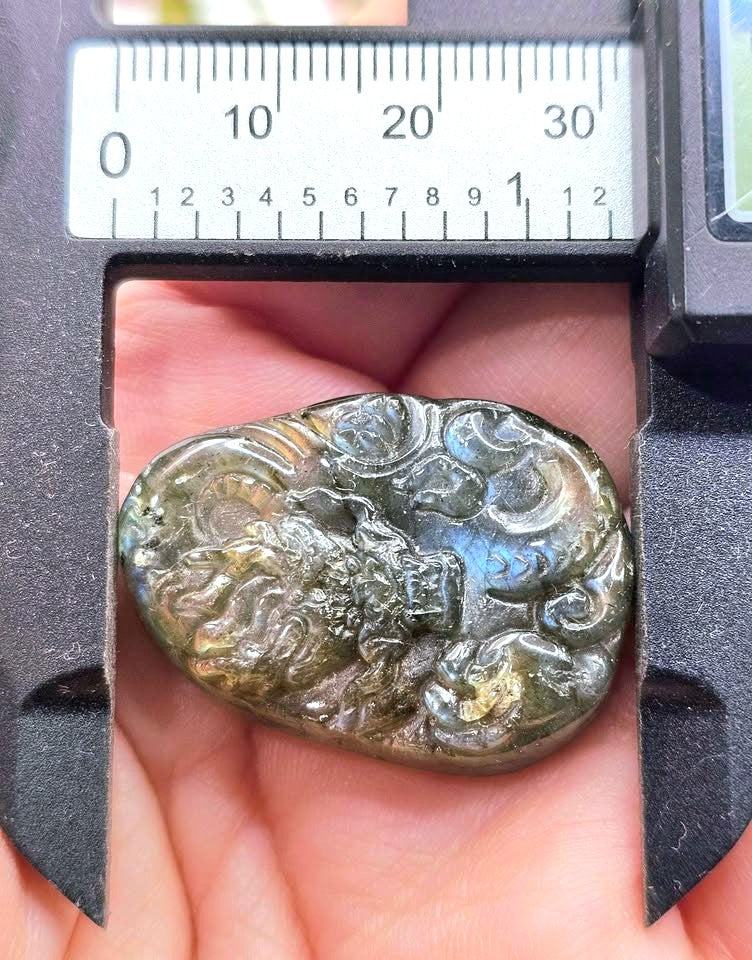 A close-up of a carved stone pendant or amulet held in a person's hand, with a caliper measuring device visible above it for scale. The stone has an embossed cloud or wave-like relief pattern and a glossy, iridescent surface showing olive, green, and blue tones.