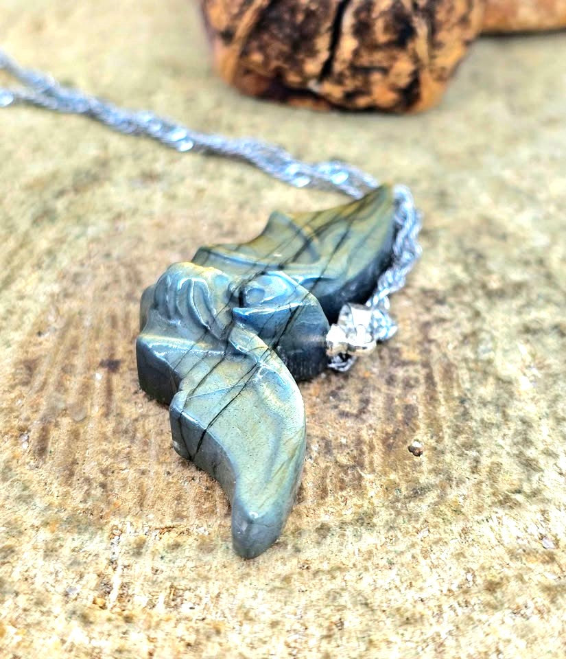 A close-up of a handmade pendant necklace featuring a carved bat-shaped piece made from layered green and blue stone with black veins, attached to a silver chain, resting on a textured tan stone surface.