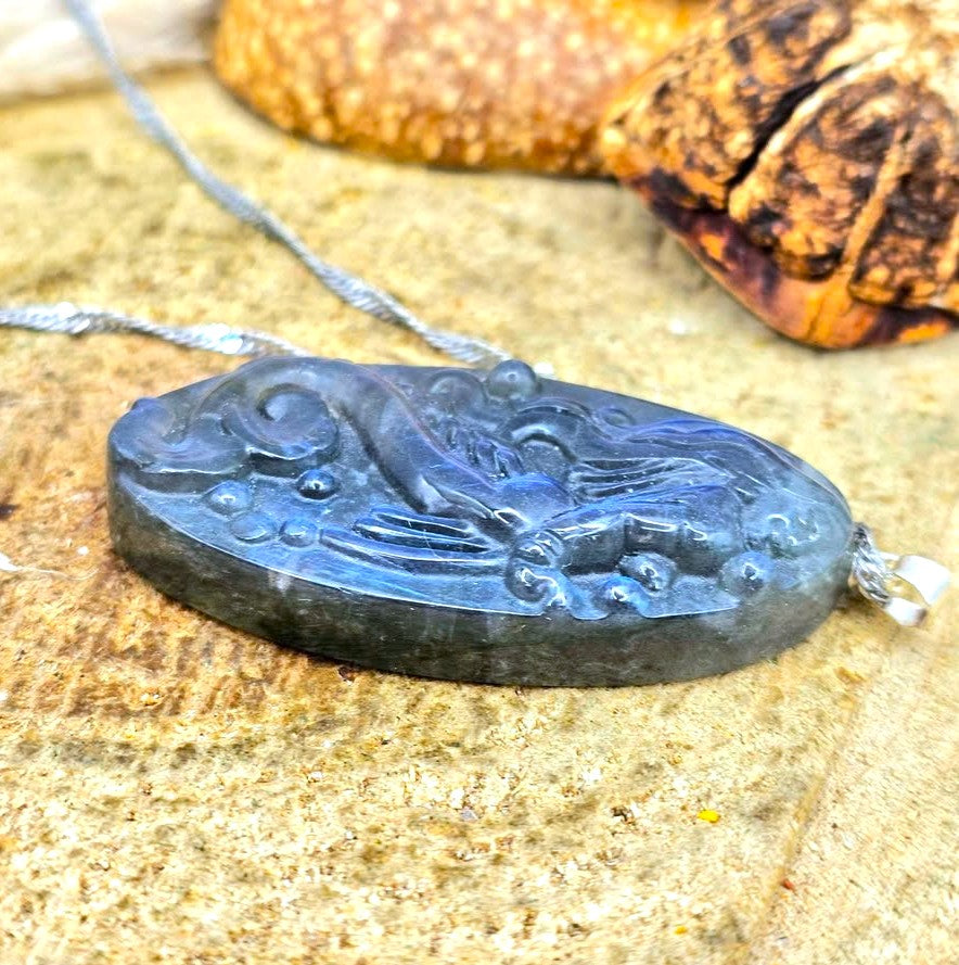 Labradorite carved pendant featuring a mermaid design, suspended from a silver chain, displayed on a sandy surface with dried grass and a small shell in the background in side view-My Aura Gemstones