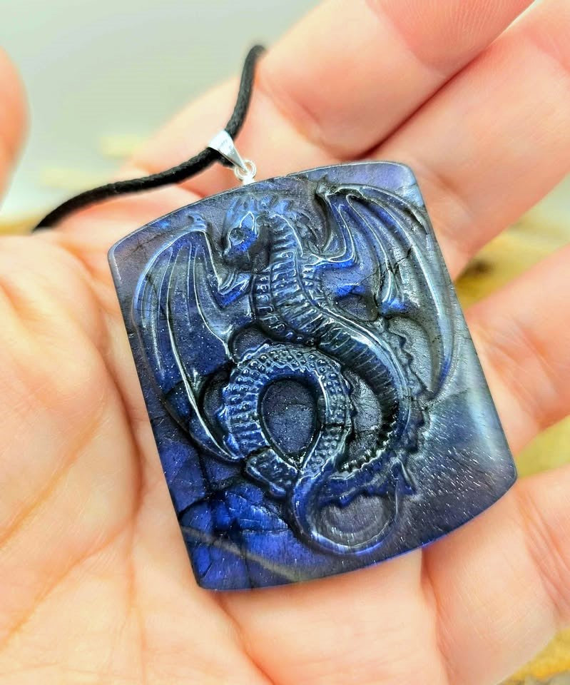 A rectangular blue labradorite pendant featuring a carved dragon design, hanging from a black cord on hand-My Aura Gemstones