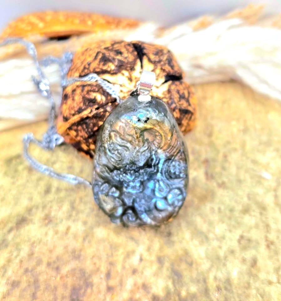 A carved stone pendant necklace with a silver-toned chain, resting on a textured tan surface with dried plant material in the background in top view-My Aura Gemstones