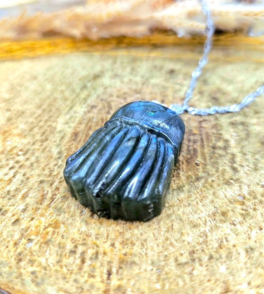 A blue iridescent jellyfish pendant necklace with a silver bail and chain, resting on a textured beige surface with dried grass and a small brown seed pod in the background at bottom view-My Aura Gemstones