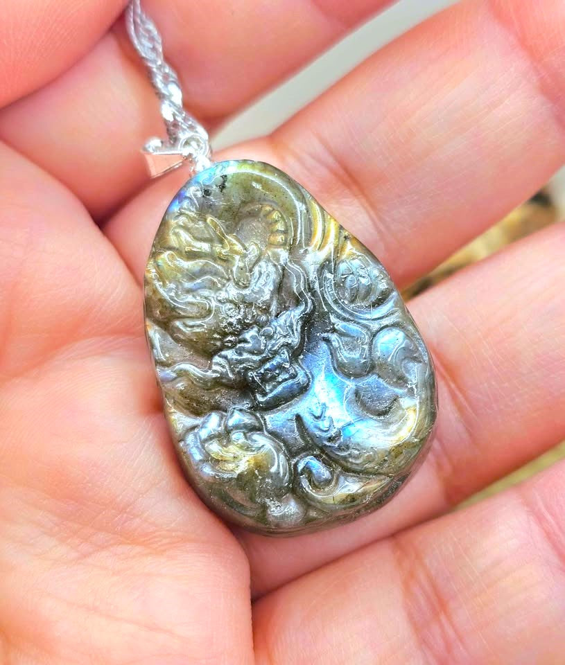A teardrop-shaped labradorite pendant necklace featuring a detailed carved dragon design, resting on hand-My Aura Gemstones