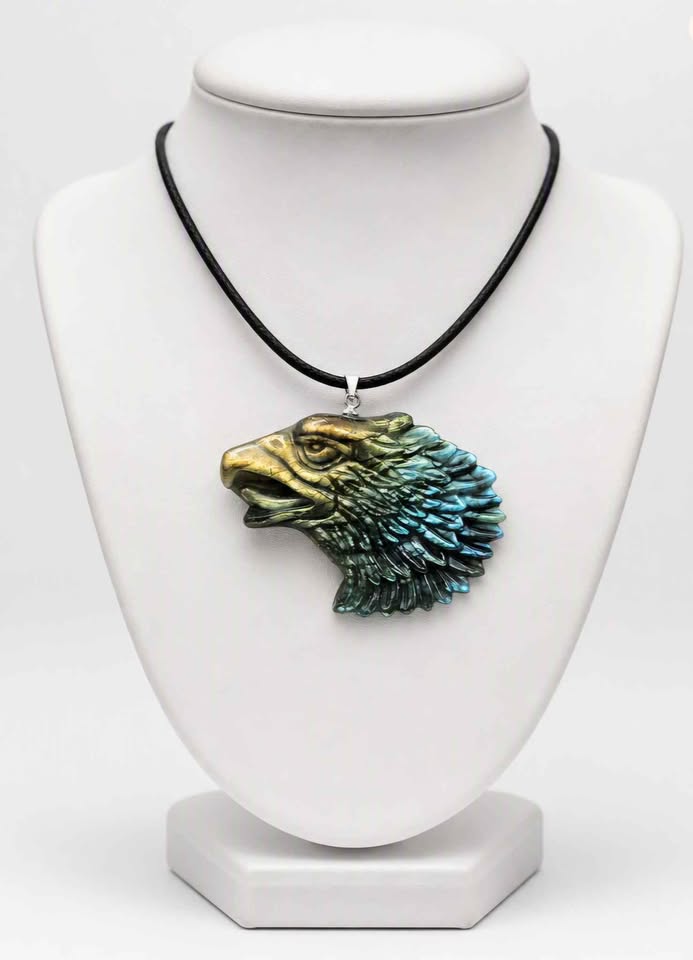 A necklace displayed on a white jewelry bust, featuring a black cord and a detailed labradorite eagle head pendant with a gold-toned face and blue-green feathered texture.