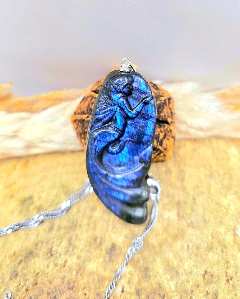 A carved labradorite pendant featuring a mermaid design, hanging from a silver chain against a rustic background of wood and dried grass.-My Aura Gemstones in top view